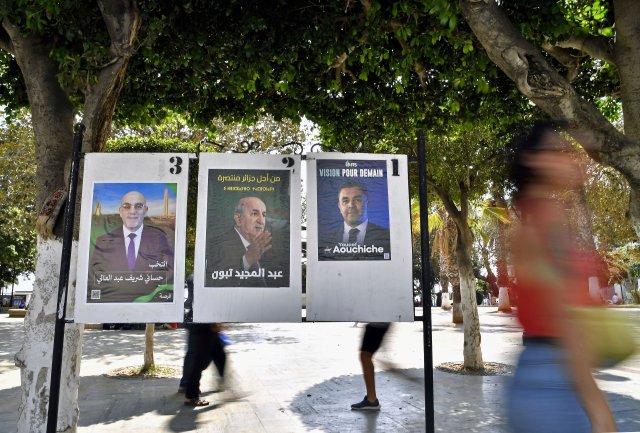 Menschen in Algier gehen an Wahlplakaten der drei Kandidaten für die Präsidentschaftswahl vorbei.