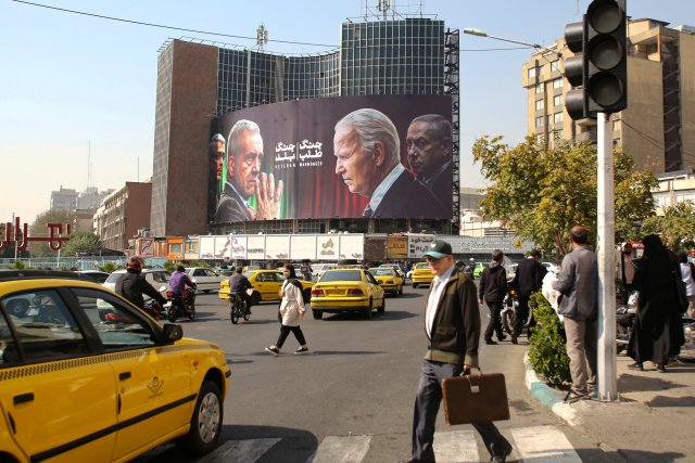 Der Normalität am Tag nach dem israelischen Vergeltungsschlag: Menschen gehen am 27. Oktober 2024 auf dem Vali-Asr-Platz in Teheran an einer Plakatwand mit Bildern des iranischen Präsidenten Masoud Pezeshkian (2.v.l.), des Generalstabschefs der Streitkräfte, Generalmajor Mohammad Bagheri (l.), des US-Präsidenten Joe Biden (2.v.r.) und des israelischen Premierministers Benjamin Netanyahu (r.) vorbei.